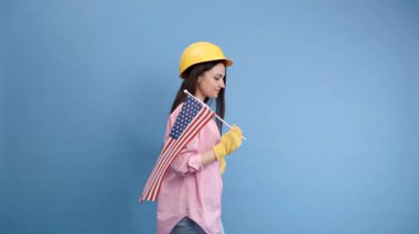 İnşaat kasklı ve eldivenli bir kız mavi arka planda Amerikan bayrağı taşıyor.