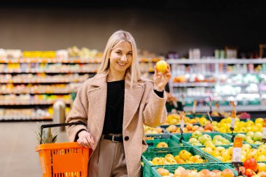 Süpermarketten alışveriş yaparken elinde elma ve alışveriş sepeti tutan gülümseyen kadın.
