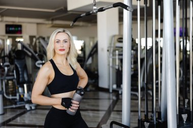 Spor salonunda kablo bağlama makinesi kullanan, elinde su şişesi tutan, siyah spor giyim ve eldiven giyen, güçlü ve kararlı görünen atletik bir kadın.
