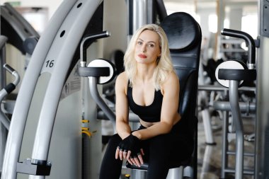 Sarışın sporcu kadın egzersiz makinesinde dinleniyor, spor aletleriyle dolu modern bir spor salonunda hak ettiği molası veriyor.