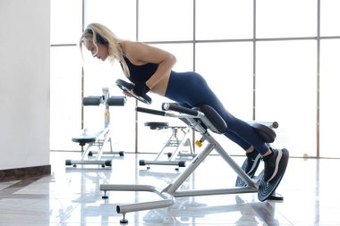 Sarışın sporcu, modern bir spor salonunda, doğal ışıkla çevrili, bir hipergenişleme makinesinde sırt kaslarını çalıştırıyor.