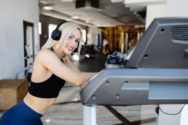 Sporcu kadın koşu bandına yaslanırken gülümsüyor, modern bir spor salonunda antrenmanının tadını çıkarıyor, vücut bulmuş spor yapıyor ve sağlıklı bir hayat sürüyor.