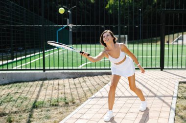Güneşli bir günde tenis oynayan genç bir kadın zarif beyaz bir spor kıyafeti içinde ustalıkla forehand vuruyor.