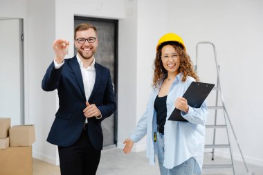 Miğferli kadın tasarımcı, elinde belgelerle odada dururken evden genç iş adamına anahtar veriyor..