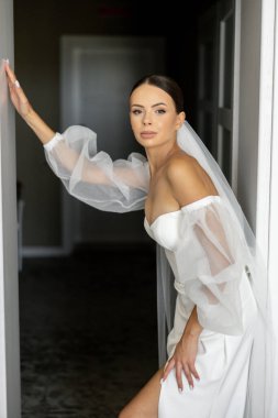 Beyaz gelinlik ve duvak giyen zarif gelin odasına giriyor ve arkasını kolluyor.