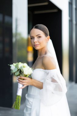 Modern binanın yanında yürüyen, beyaz gelinlik ve duvak giyen, gelinlik modasını sergileyen zarif gelin.