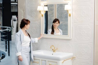 Açık gri takım elbiseli bir kadın modern bir galeride duruyor, çerçevenin sağ tarafına bakıyor. Ön planda büyük, beyaz, serbest bir küvet var. Kadının sağ eli cebinde, ve sol eli de cebinde duruyor.