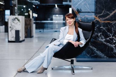 Açık mavi takım elbiseli bir kadın siyah deri döner bir sandalyede oturur. Arka plan mermer duvarlı modern bir ofis alanı. Kadın yan tarafa bakıyor..