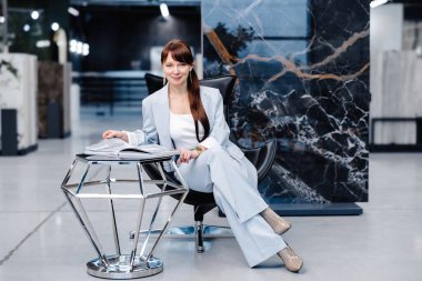 Açık mavi takım elbiseli bir kadın siyah deri bir sandalyede oturuyor ve kucağında bir dergi var. Topuklu giyiyor ve kameraya bakıyor. Önünde geometrik gümüş bir masa duruyor. Arka plan mermer bir duvar..