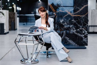 Açık mavi takım elbiseli bir kadın siyah deri bir sandalyede oturuyor ve kucağında bir dergi var. Topuklu giyiyor ve kameraya bakıyor. Önünde geometrik gümüş bir masa duruyor. Arka plan mermer bir duvar..