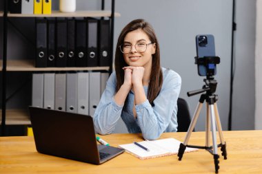 Video konferansı ve eğitim. İnternet uygulaması kullanarak çeşitli öğrencilerin uzaktan öğrendiği bir sınıf. Dişi öğretmen dizüstü bilgisayarla ders veriyor, cep telefonunu tripoda kaydediyor..