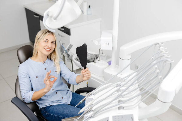 Young woman showing ok sign while holding dental mirror and sitting in dentist chair, satisfied with treatment result
