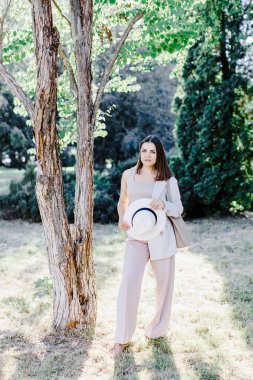 Beyaz, kendine güvenen, takım elbiseli, parkta yürüyen iş kadınının vücut portresi. Şık takım elbiseli ve beyaz şapkalı güzel bir kadın parktaki ağacın yanında duruyor.