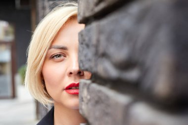 Beyaz bir dış duvara yaslanmış, kenar mahallede fotokopi alanı olan ve kameraya bakan sevimli genç bir kadın.