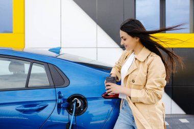 Küçük bir kamu istasyonundan şarj olan mutlu genç bir kadın, elektrikli arabanın yanında duruyor, kahve içiyor..