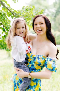 Genç ve güzel anne, sevimli kızına sarılıyor, onu kollarına alıyor, parkta bir ağacın yanında duruyor..