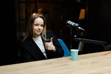 Genç bayan radyo sunucusu profesyonel bir stüdyoda podcast kaydederken başparmak işareti yapıyor.