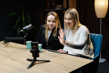 Profesyonel bir stüdyoda canlı yayın yapan iki kadın içerik yaratıcısı kameraya el sallıyor.