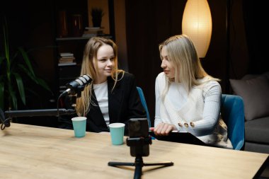 Profesyonel mikrofon ve akıllı telefon kullanarak canlı yayında gülümseyen ve kaydeden iki kadın podcaster.