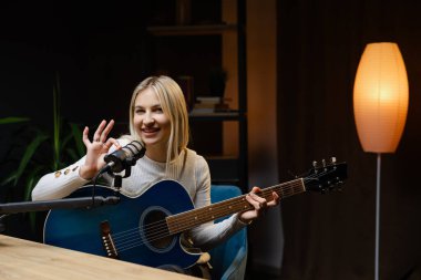 Müzisyen, profesyonel mikrofon ve mavi akustik gitar kullanarak ev stüdyosunda şarkı kaydediyor.