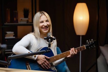 Gülümseyen bayan müzisyen bir stüdyoda şarkı söylerken akustik gitar çalıyor.