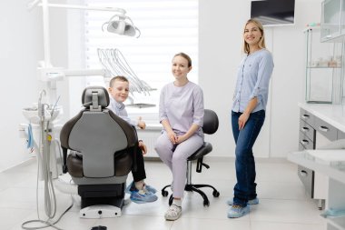 Diş hekimi bir anne ve oğluyla diş hijyeni hakkında konuşuyor, diş sağlığı kliniğinde oturuyor ve ayakta duruyor.