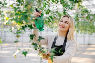 Gülümseyen kadın bahçıvan verimli sarmaşık yapraklarına su püskürterek canlı bir seradaki bitkileri besliyor, sağlıklı büyümeyi teşvik ediyor.