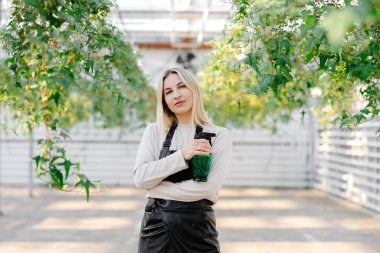 Kendine güvenen bahçıvan elinde sprey şişesiyle serada dikiliyor, çiçeklerle ilgileniyor ve gülümsüyor.