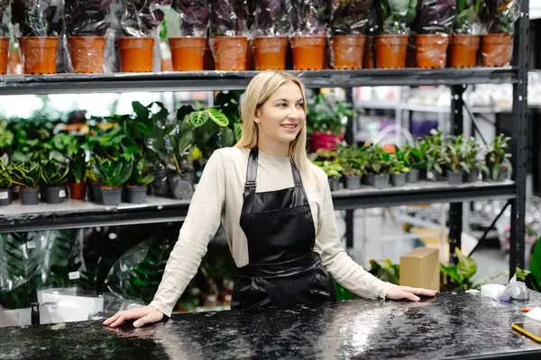 Çiçekçi dükkanında siyah önlüklü genç bir kadın dikiliyor, etrafı bitkilerle çevrili.