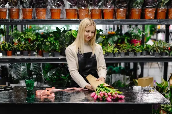 Çiçekçide pembe lale buketi düzenleyen, kahverengi kağıtla saran, teslimat ya da müşteri alımına hazırlanan bir çiçekçi.