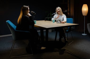 İki mutlu kadın podcaster yeni bir bölüm kaydediyor. Profesyonel bir kayıt stüdyosunda konuşuyor ve gülüyorlar.