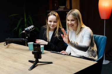 Profesyonel bir stüdyoda canlı yayın yapan iki kadın içerik yaratıcısı kameraya el sallıyor.