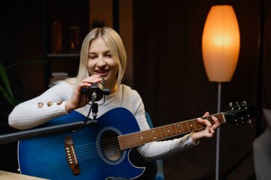 Müzisyen, profesyonel mikrofon ve mavi akustik gitar kullanarak ev stüdyosunda şarkı kaydediyor.