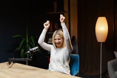 Bir stüdyoda podcast çekerken başarıyı kutlayan genç bir kadın heyecanla ve başarıyla kollarını kaldırıyor.