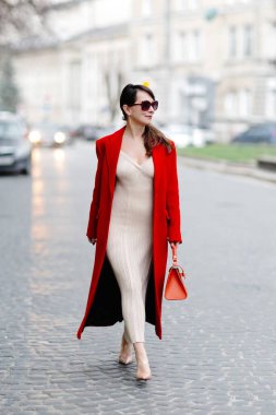 Çarpıcı kırmızı ceketli ve şık güneş gözlüklü bir iş kadını şehir manzarasında kaldırım taşı caddesinde güvenle yürüyor.