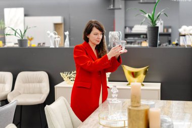 Kırmızı ceketli bir kadın lüks bir mobilya mağazasında alışveriş yaparken yemek odası için zarif kristal bardaklar seçiyor.