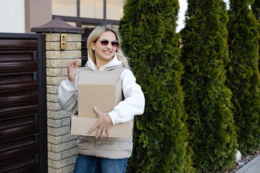 Elinde karton kutular ve evin girişinde güneş gözlüğü olan gülümseyen teslimatçı kadın.