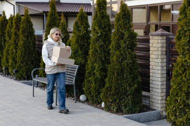 Genç bir kadın, güneşli bir bahar gününün tadını çıkarırken, meskun bir mahallede kaldırımda yürürken iki karton kutu taşıyor.