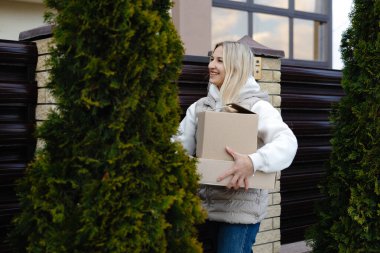 İki karton kutu taşıyan gülümseyen kadın evinin bahçesine giriyor ve iki thuja ağacının arasından geçiyor.