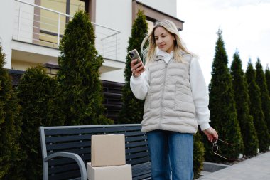 Dışarıdaki bankta karton kutuların yanında siparişini beklerken cep telefonu kullanan genç bir kadın.