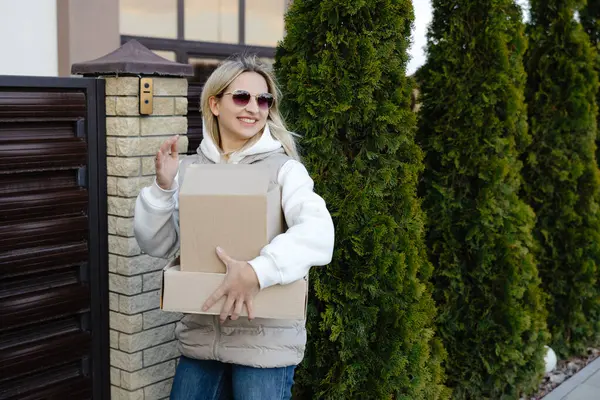 Elinde karton kutular ve evin girişinde güneş gözlüğü olan gülümseyen teslimatçı kadın.