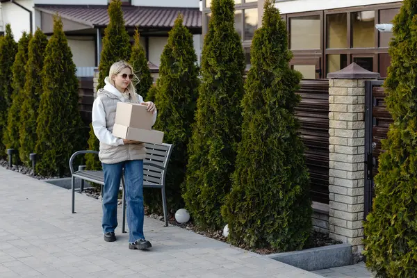 Genç bir kadın, güneşli bir bahar gününün tadını çıkarırken, meskun bir mahallede kaldırımda yürürken iki karton kutu taşıyor.