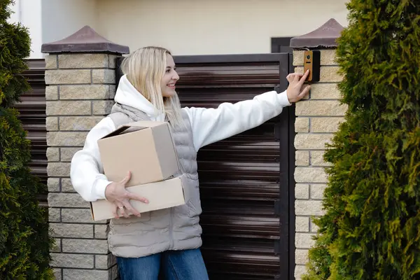 Elinde karton kutular ve evin girişinde güneş gözlüğü olan gülümseyen teslimatçı kadın.