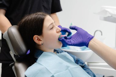 Diş hekimi, diş çürüklerini önlemek için hastanın dişlerine tükürük püskürtücü tutarak diş macunu sürüyor.