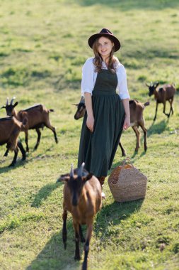 Şapka takan genç bir kadın, güneşli bir tarlada keçiyi nazikçe okşuyor. Etrafı hasır sepet ve canlı yeşillikle çevrili.