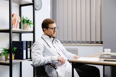 A young male doctor sits at his workplace and feels tired from work. A doctor takes a break from his work day because of pain.
