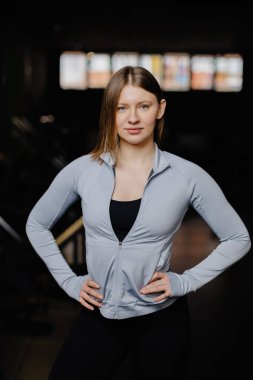 Spor salonunda elleri kalçalarının üzerinde duran genç bayan sporcu yoğun bir idmandan sonra özgüven ve kararlılık sergiliyor.