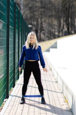 Formda bir kadın, güneşli bir açık havada direniş bandıyla çömelme hareketleri yapıyor. Egzersiz sırasında gücünü ve bağlılığını gösteriyor.