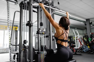 Siyah takım elbiseli kaslı atletik bayan vücut geliştirici spor salonunda spor simülatörüne çekildi. Sırt kasları çok gergin.