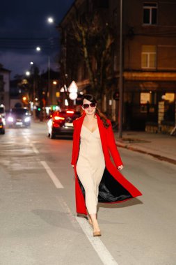 Şık kırmızı paltolu ve şık güneş gözlüklü zarif bir kadın gece şehir ışıklarıyla çevrili canlı bir caddede güvenle yürüyor.